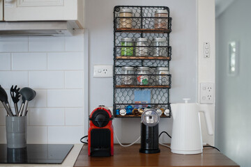 kitchen bench with coffee machine, milk spinner and kette. on the wall are power points with nothing plugged in, and a shelf of jars of tea, coffee and sugar, appliance