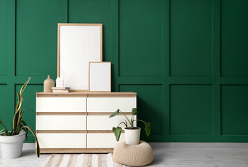 Chest of drawers with houseplants and blank posters near green wall in room