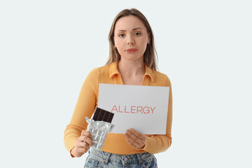 Young woman holding paper with word ALLERGY and chocolate bar on white background