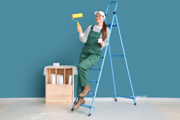 Female worker with paint roller and stepladder near blue wall