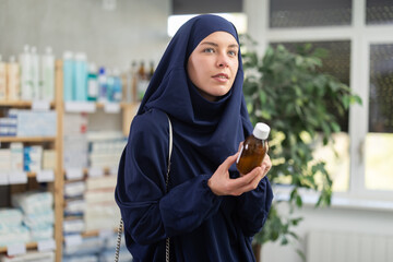 Thoughtful young Muslim woman wearing navy blue hijab and abaya holding and reading label of cough...