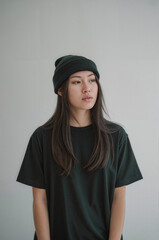A portrait photograph of an Asian woman wearing a dark green beanie and an oversized black t-shirt against a plain light gray background.