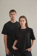 A minimalist studio portrait of two young people standing against a plain off-white wall. Both are wearing plain black t-shirts and dark pants. 