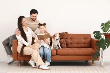 Cute little girl with her parents and Beagle dog reading book on sofa at home