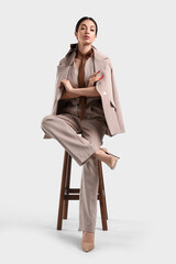 Stylish elegant young woman in beige suit sitting on stool against white background