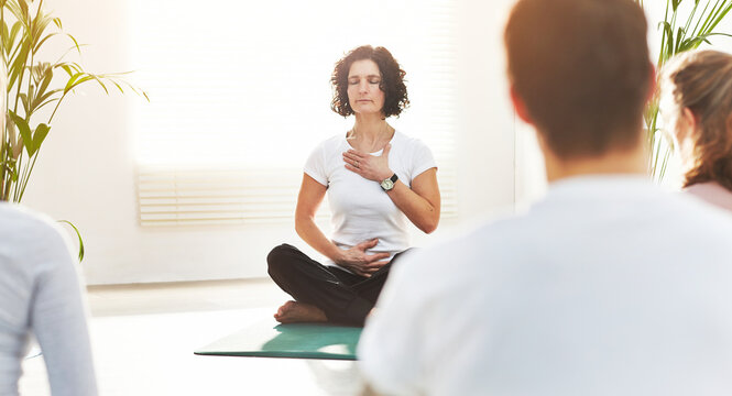 Yoga instructor training people in a fitness class in studio. Yogi teaching pranayama breathing techniques with hands on chest and belly. Practicing mindful meditation for zen energy in lotus pose