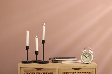 Candlesticks with burning candles, alarm clock and books on wooden cabinet near light brown wall...