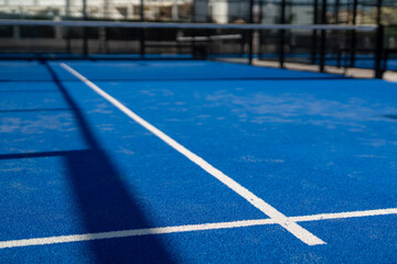 White court line on blue synthetic padel court surface, close-up perspective with net and shadows on a sunny outdoor sports field