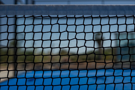 Close-up of black padel court net with hexagonal mesh pattern, blue sports field and blurred background under sunlight