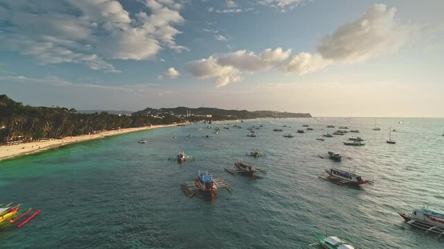 Aerial drone view of Cagban Beach in Boracay Island, Philippines, showcasing numerous traditional banca boats dotting the vibrant turquoise waters under a picturesque sunset sky