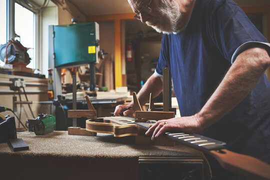 Creative guitar maker designing an artistic wooden musical instrument alone inside his professional workshop. Skilled, serious and smart mature luthier craftsman building and repairing instruments - Powered by Adobe