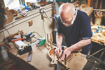 Skilled, expert and professional carpenter creating a guitar with wood in a workshop. Senior male craftsman, handyman or artisan making a handmade wooden instrument in a woodwork and carpentry shop