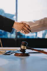 Two businesspeople are shaking hands in a law office with a scale and a gavel on the table.