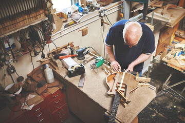 Creative and skilled carpenter making a guitar out of wood in a workshop. Above view of senior...