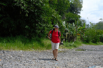 Side view: A woman walks along on a rocky path surrounded by green trees under a bright; clear sky
