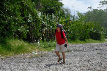 Side view: A woman walks along on a rocky path surrounded by green trees under a bright; clear sky