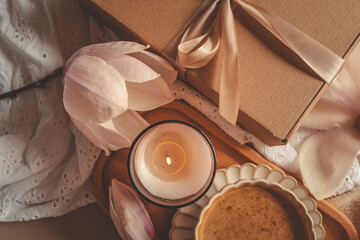 Lit candle in a glass jar with soft magnolia flowers, a gift box, and a coffee cup in warm pastel light on white lace fabric
