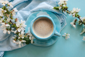 A cup of coffee with foam in a blue cup with white cloud design, surrounded by white spring blossoms, a soft blue fabric, and an open book, evoking a serene and cozy atmosphere