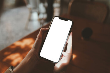 Hands holding smartphone with blank white screen in cozy indoor setting, relaxed atmosphere with wooden furniture, digital technology and communication concept.
