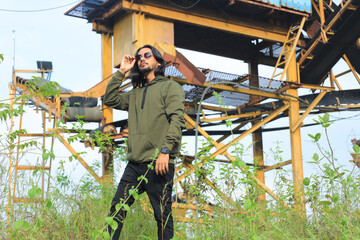 A young man with long hair and a beard stands confidently on a rocky lakeside shore, wearing a green hoodie, black jeans, and white sneakers