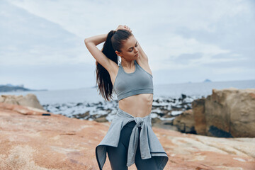 A young woman of mixed descent stretches on rocky coastline, wearing a gray sports bra and black...
