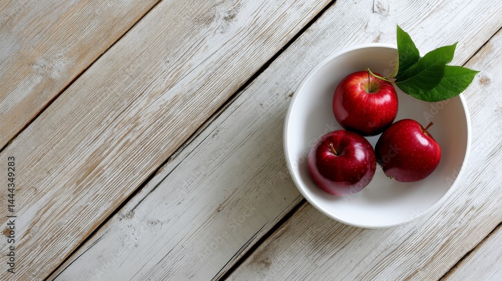 Poster A top-down view highlights three ripe red apples with a green leaf in a simple white bowl, elegantly positioned on a light wooden backdrop