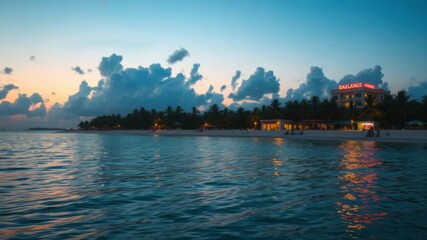 Naklejka premium A picturesque beach scene at sunset with a hotel illuminated against a vibrant sky and calm ocean waters reflecting the lights.