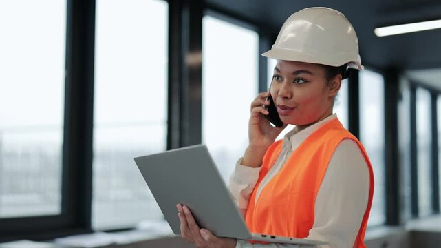 Confident female engineer in an orange vest and hard hat reviews blueprints on a laptop. She discusses plans in a phone call from a modern office building.
