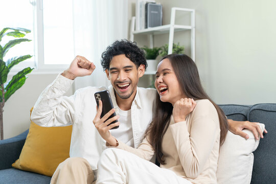 Relax and happiness lifestyle at home concept. Young couple sitting on sofa in living room looking at smartphone smile on face in cozy atmosphere.