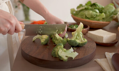 Close-up of a person slicing fresh broccoli on a wooden board, surrounded by tofu and leafy greens, ideal for healthy cooking themes.