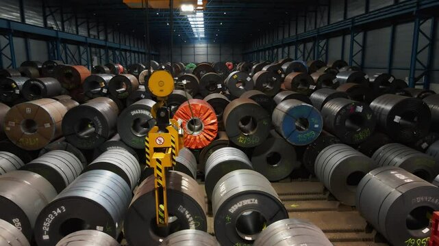 A large crane grabs a heavy steel coil and picks it up in a large warehouse