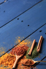 A collection of colorful spices, including cinnamon sticks, red pepper flakes, green herbs, and turmeric powder, are scattered on a blue wooden surface with spoons.
