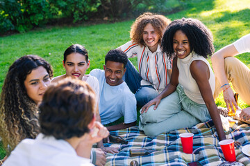 Group of diverse young people having fun, eating and drinking at a picnic in a park, enjoying summer holidays together