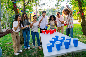 Cheerful multi ethnic friends enjoying a lively beer pong game at a vibrant park party, celebrating friendship and summer fun together