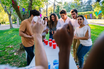 Group of multi ethnic friends enjoying a sunny day outdoors while playing beer pong in a city park,...