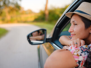 Traveler woman in red ethnic dress and hat drives car through scenic countryside of Croatia in summer. Surrounded by lush fields and forests, she enjoys peaceful and adventurous road trip