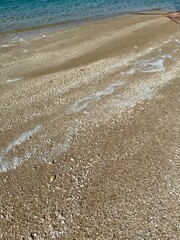 Low-angle shoreline view showing foamy wave trails on coarse sand leading to calm turquoise water, with a red fishing net barely visible in the top corner.