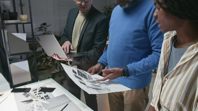 Side tilt shot of group of three diverse developers using laptop and technical sketches, brainstorming ideas about enhancing sustainable technologies and implementing renewable solutions in life