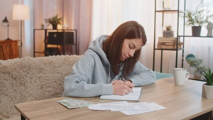 Focused young Caucasian woman making shopping list in notebook sitting at table on sofa looking at receipts and counting money in living room at home. Girl preparing notes for financial cost or budget