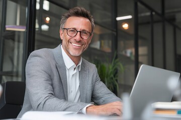 Happy businessman with laptop smiling at the camera in the office. The man is sitting at a desk using a computer, wearing glasses and a grey suit