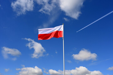 The polish flag flutters in the wind an aeroplane flies past at high altitude with a contrail