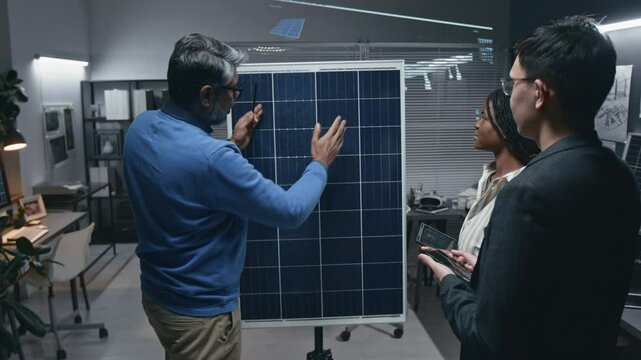 Arc shot of senior male Indian engineer-mentor explaining work of solar panel photovoltaic cells to two diverse trainees in modern sustainable technology office