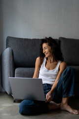 Naklejka premium Happy young freelance woman working from home with laptop, sitting on the floor in casual clothes, smiling and enjoying remote work in cozy relaxed atmosphere.