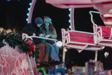 Naklejka premium A couple enjoys a festive amusement ride decorated with holiday lights and ornaments during winter. The setting evokes warmth, celebration, and joyous moments shared on a chilly evening.