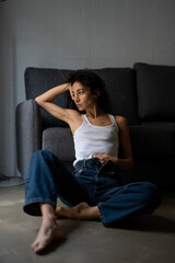 Obraz premium Thoughtful woman in white tank top and jeans sitting barefoot on the floor beside a dark gray sofa, resting her head on hand and gazing out in soft natural light.