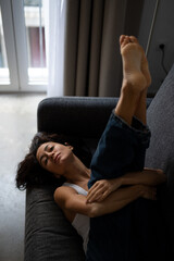 Fototapeta premium Natural light portrait of a woman resting barefoot on a grey sofa, lying on her back with legs raised, wearing jeans and a white tank top, serene indoor atmosphere.