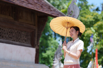 Smiling thai woman in traditional clothing holding a parasol while standing near a wooden temple in...