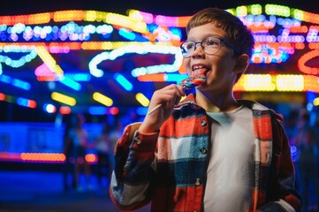 A cute caucasian boy eating a colorful lollipop standing against the background of a carousel in...