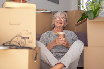 Happy senior woman involved in moving house sitting among cardboard boxes having a pause, concept of relocating, retirement, new life, buying, renting, apartment, house