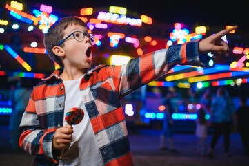 A cute caucasian boy eating a colorful lollipop standing against the background of a carousel in the evening at an amusement park or circus © Serhii